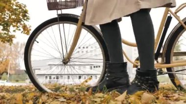 Woman walks with a bicycle in the autumn park with fallen leaves, legs in black boots close-up side view