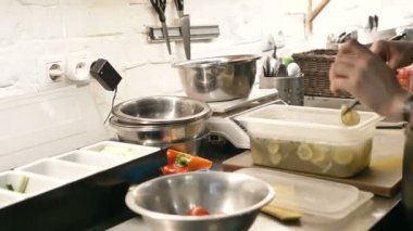 young person cuts fresh cucumber and puts slices into metal bowl on digital scales at table in light restaurant kitchen closeup