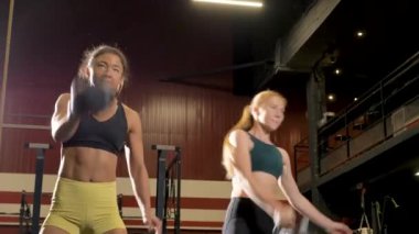Brunette and ginger girl friends wearing sport tops shorts and leggings do squats and weightlifting barbells actively in fitness centre