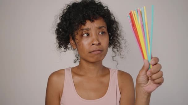 アフリカ系アメリカ人女性は灰色の背景に立っているプラスチックと有機藁を比較します 若い主婦は環境に優しいストローを選びます — ストック動画