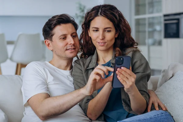 Excited married woman with long curly hair and brunet man smile happily reading good news in internet via smartphone sitting on sofa