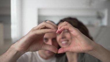 Happy married couple with dark hair and broad smiles kisses and shows heart sign with hands taking photos against blurry kitchen closeup