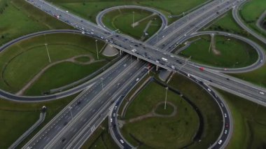 Cars drive along huge multi-level autobahn traffic intersection among green meadows against distant city in mist aerial panorama