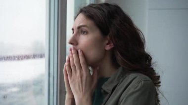 Brunette woman with curly hairstyle looks in shock out of window at crime scene at police flashing red lights with raindrops on glass