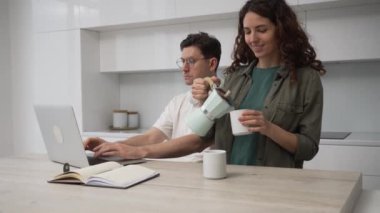 Man with headphones on neck frowns working distantly via laptop and brunette wife pours coffee into cups to cheer up husband smiling and kissing