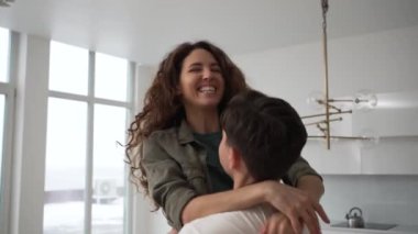 Young brunet man catches holding in arms and spins curly wife in shirt and jeans hugging tightly and laughing thankfully in kitchen closeup