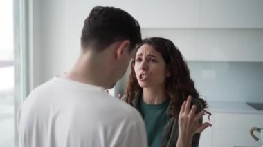 Married curly-haired woman quarrels angrily gesticulating and shouting with brunet husband over family problems standing in kitchen closeup