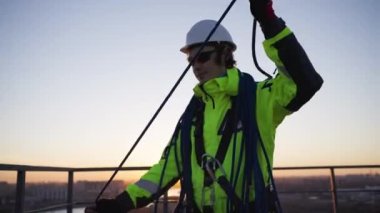 Male industrial climber in a hard hat and overalls collects a rope after a work shift on the roof of a building against the sunset. The engineer performed high-rise work on the facade of the building.