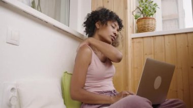 African American woman works on laptop kneading neck on balcony. Young freelancer leans on pillow looking in electronic device slow motion