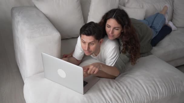 暗い髪を持つ幸せな夫婦はソファに横たわっていると笑顔広くラップトップ経由で改装されたアパートのためのオンライン新しい家具を購入 — ストック動画