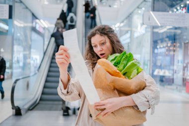 Şokta olan bir kadın, alışveriş merkezindeki bir süpermarketten alınan kağıt çekle yürüyen merdiven arkaplanını karşılaştırıyor ve elinde içinde yeni ürünler olan bir paket tutuyor.