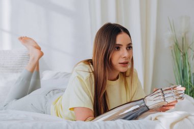 Biyonik protez kollu bir kadın blogcu, bir internet yayınını yönetiyor veya bir konuyu kaydediyor veya güneşli bir günde rahat ev kıyafetleri içinde bir yatakta yatarken video aracılığıyla arkadaşlarıyla iletişim kuruyor..