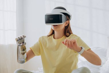 Şık bir biyonik protez kolu ve sanal gerçeklik gözlüklü neşeli bir kadın evdeki yatakta oturmuş video oyunu oynuyor.