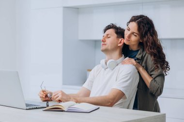 Wife is massaging the shoulders of her tired husband, a freelancer programmer in glasses and headphones, working on a laptop at home in the kitchen. Husband took a break from work.