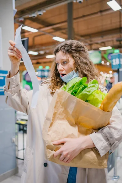 슈퍼마켓에서 의료용 마스크를 여자가 수표를 이봉지에 식료품을 놀라서 가격을 바라본다 — 스톡 사진