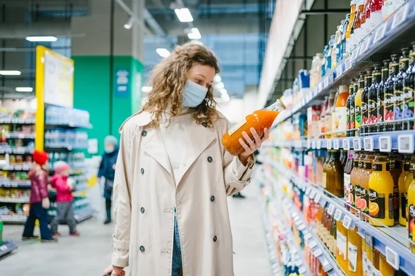 Young Curly Woman Medical Mask Beige Raincoat Chooses Juice Glass — Stockfoto