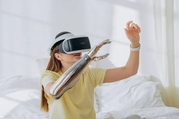 Joyful woman with stylish bionic prosthesis arm and virtual reality goggles plays video game sitting on bed in room at home