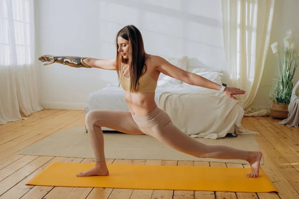 Cheerful woman with metal bionic prosthesis arm does stretching exercises on floor near bed in sunny room closeup