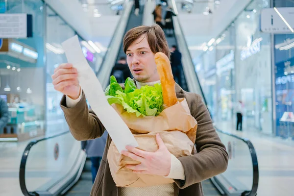 コートの男は ショッピングモールでバゲットとレタスと紙のチェックと袋を保持し 食料品の高価格にショックを受けています ロイヤリティフリーのストック写真