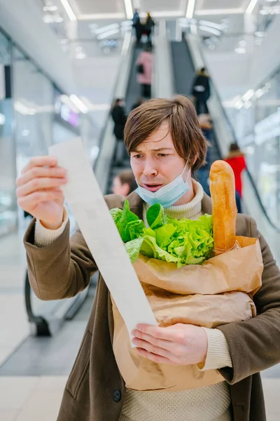 医療マスクの男はスーパーマーケットでのチェックで恐怖を見て 経済危機のために食料品 価格の上昇とインフレと紙袋を保持しています ストックフォト