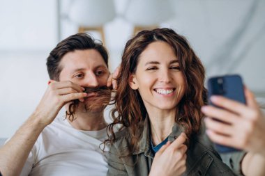 Gülümseyen esmer kadın ve kısa saçlı genç adam fotoğraf ve videolar için bıyık şeklinde kıvırcık saçlı poz veriyorlar.