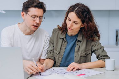 Couple sits in kitchen drinks coffee for breakfast and talks about drawings of apartment renovation making notes in laptop and notebook