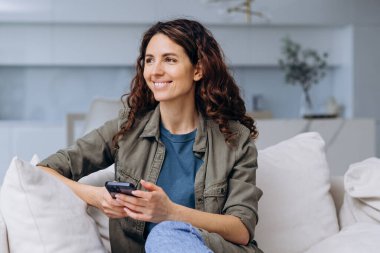 Evdeki kanepede oturup pencereden dışarı bakarken elinde akıllı telefonuyla gülümseyen mutlu genç bir kadın güzel bir mesaj aldı ya da tatille ilgili rüyalar gördü.