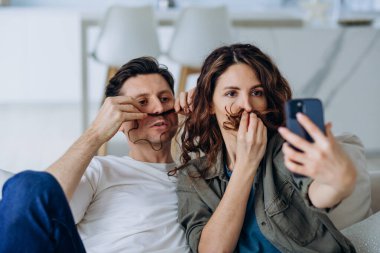 Gülümseyen esmer kadın ve kısa saçlı genç adam fotoğraf ve videolar için bıyık şeklinde kıvırcık saçlı poz veriyorlar.