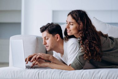 Newly married woman and man working on laptop start choosing decorations for renovated flat on sofa in living room and discuss variants smiling
