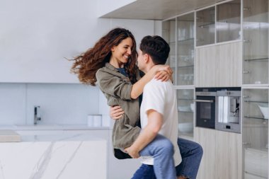 Young brunet man catches holding in arms and spins curly wife in shirt and jeans hugging tightly and laughing thankfully in kitchen closeup