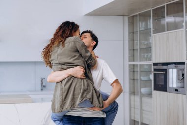 Young brunet man catches holding in arms and spins curly wife in shirt and jeans hugging tightly and laughing thankfully in kitchen closeup