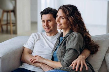 Happy newlyweds with dark hair sit on comfortable sofa in living room and discuss plans for future smiling broadly closeup