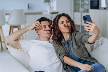 Laughing husband puts young wife long curly locks as theatrical mustache and poses against kitchen for funny videos and selfies closeup