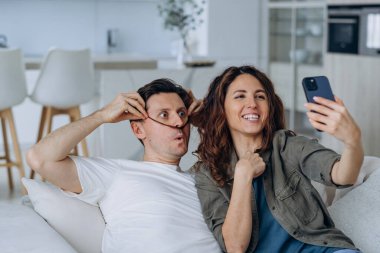 Cheerful in love couple taking a selfie on the phone while sitting on the couch together, making funny mustaches out of their hair and taking pictures