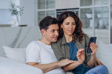 Excited married woman with long curly hair and brunet man smile happily reading good news in internet via smartphone sitting on sofa