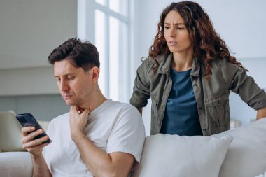 Young woman looks out over husband shoulder at phone screen and starts quarreling accusing man in cheating in living room clipart