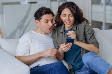Excited married woman with long curly hair and brunet man smile happily reading good news in internet via smartphone sitting on sofa