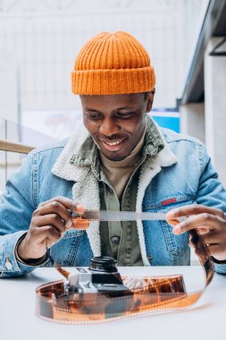 Kot ceketli gülümseyen Afro-Amerikan fotoğrafçı eski filmlerdeki fotoğraflara bakıyor.