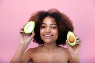 Genç ve güzel bir Afro-Amerikan kadının portresi gür saçlı pembe arka planda kesilmiş bir avokado tutuyor ve kameraya bakıyor.
