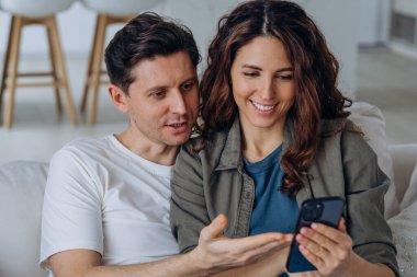 Genç esmer koca ve kıvırcık karı sosyal medyadan geniş bir gülümsemeyle düğün fotoğraflarını tartışıyor ve kanepeye yakın oturuyorlar.