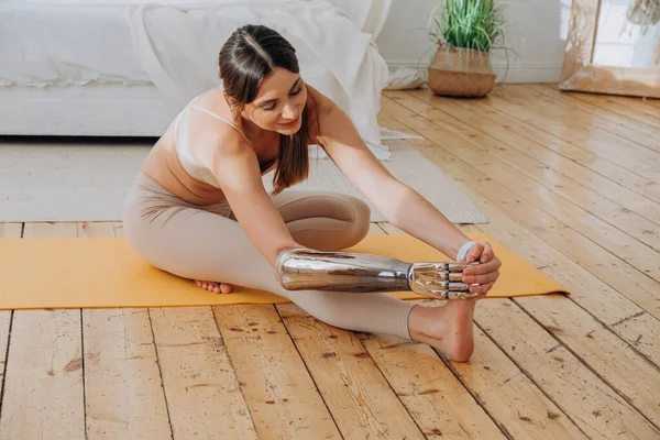 Cheerful woman with metal bionic prosthesis arm does stretching exercises on floor near bed in sunny room closeup