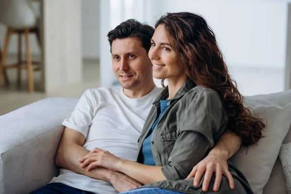 Happy newlyweds with dark hair sit on comfortable sofa in living room and discuss plans for future smiling broadly closeup