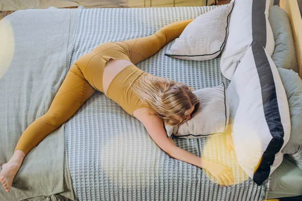Woman with blonde long hair in yellow top and leggings does stretching and split lying on bed covered with blanket and hugging pillow