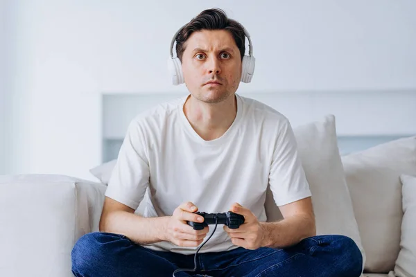 Excited man in wireless headphones sits on sofa in living room plays non-stop catch-up game with controller and loses illuminated by screen light