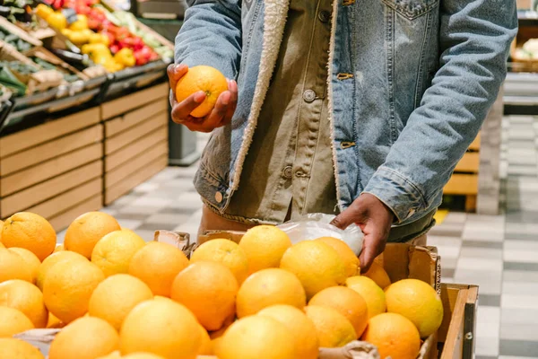 Young African-American customer in warm jacket with protective mask takes fresh orange fruits from crate in light supermarket