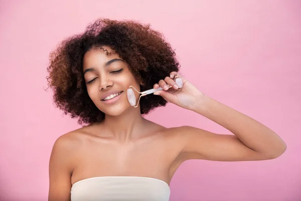 Portrait of a young well-groomed dark-skinned woman with lush hair, doing facial massage with the help of a roller with closed eyes on a pink background close-up, care and skin care
