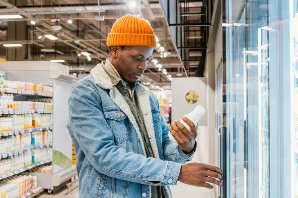 Positive African-American guy in denim jacket and orange hat takes milk bottle from fridge case in modern supermarket side view