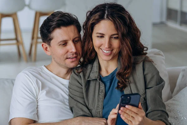 Genç esmer koca ve kıvırcık karı sosyal medyadan geniş bir gülümsemeyle düğün fotoğraflarını tartışıyor ve kanepeye yakın oturuyorlar.