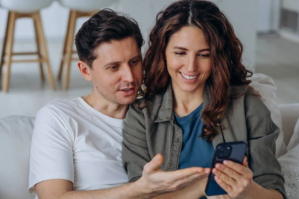Genç esmer koca ve kıvırcık karı sosyal medyadan geniş bir gülümsemeyle düğün fotoğraflarını tartışıyor ve kanepeye yakın oturuyorlar.