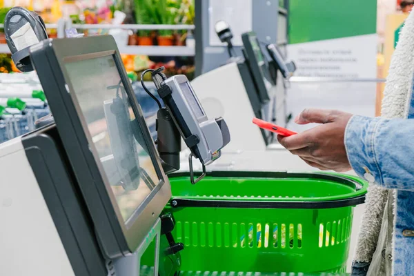 暖かいデニムジャケットの黒人男性はスーパーマーケットの近くのビューでセルフチェックアウトポイントで購入するためにスマートフォンを使用しています ストック画像